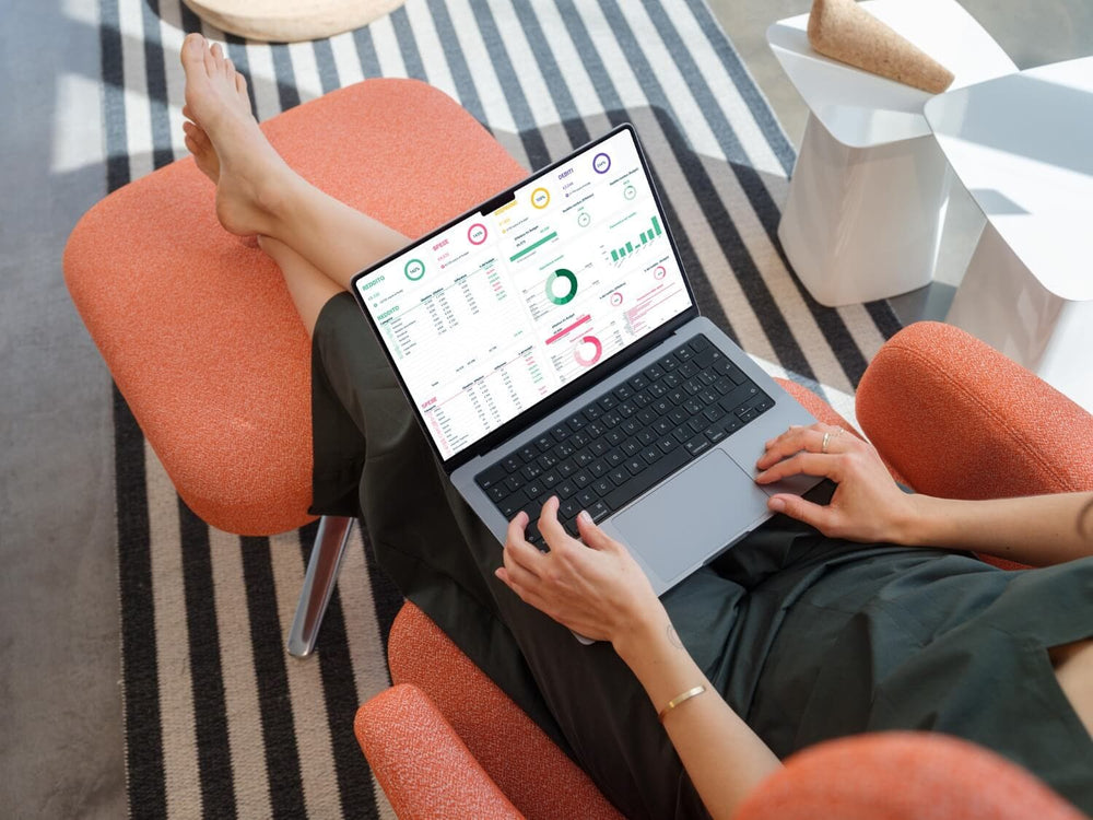 Donna rilassata su poltrona arancione mentre controlla il proprio budget mensile con un template Excel sul laptop. Schermata con grafici di spese, reddito e risparmi.